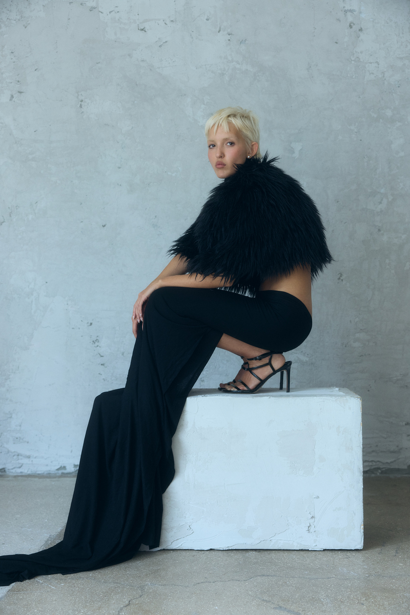 Editorial fashion portrait by Josh Eskridge of a short-haired blonde model wearing a black designer gown and feathered cape against a handmade concrete-textured wall at Know Good Studio in Miami.