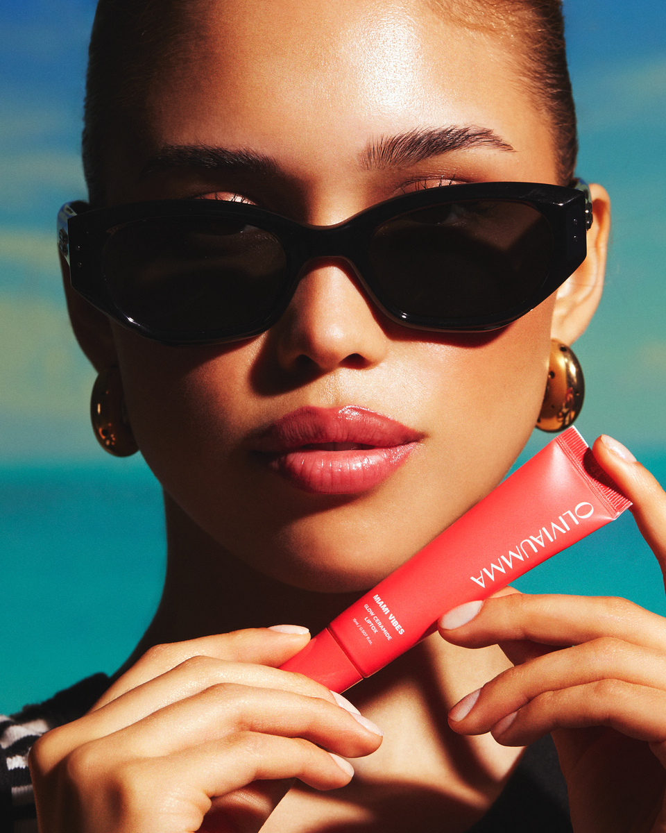 Model in black sunglasses holding Olivia Umma Miami Vibes lip product against a turquoise background.